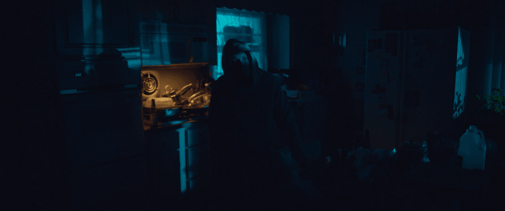 in a shadowy kitchen, a hooded figure steps into a pool of light