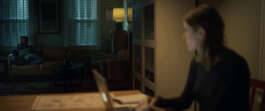 a woman works at a table while a man watches TV across the room