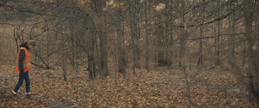 a woman in an orange vest walks though a forrest in the fall